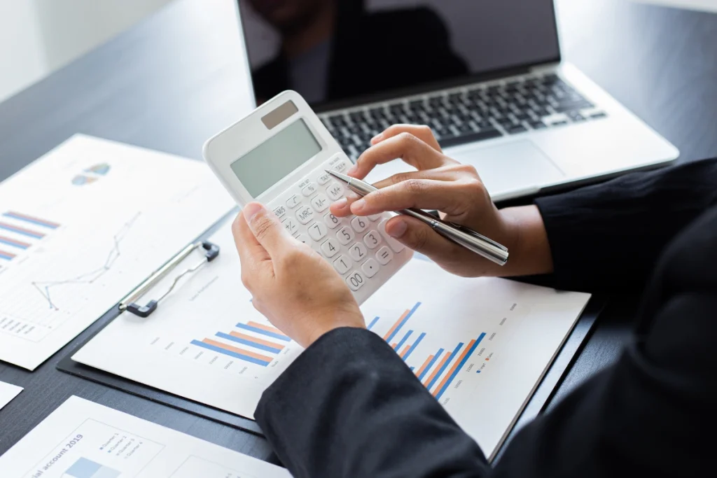 Businessperson using a white calculator while reviewing charts and graphs on paper, representing financial planning or budgeting associated with POTS Line Replacement solutions.